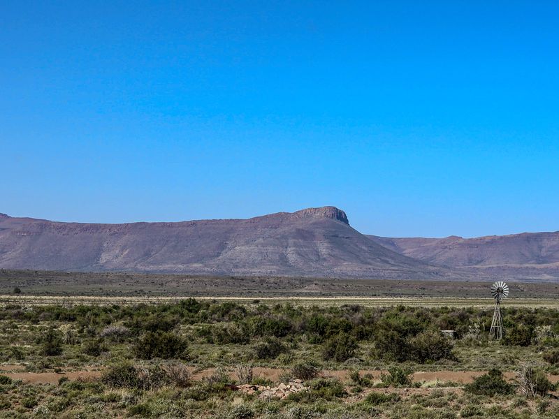 Expansive landscape view featuring a traditional farm windmill set against a backdrop of rugged mountains and open veld.