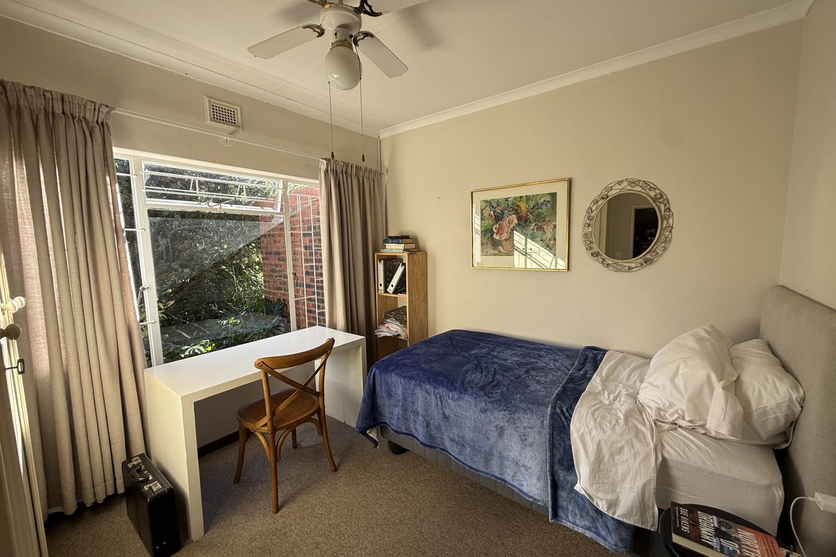 A functional bedroom featuring carpet flooring, a ceiling fan, and a large steel-framed window overlooking the garden area.