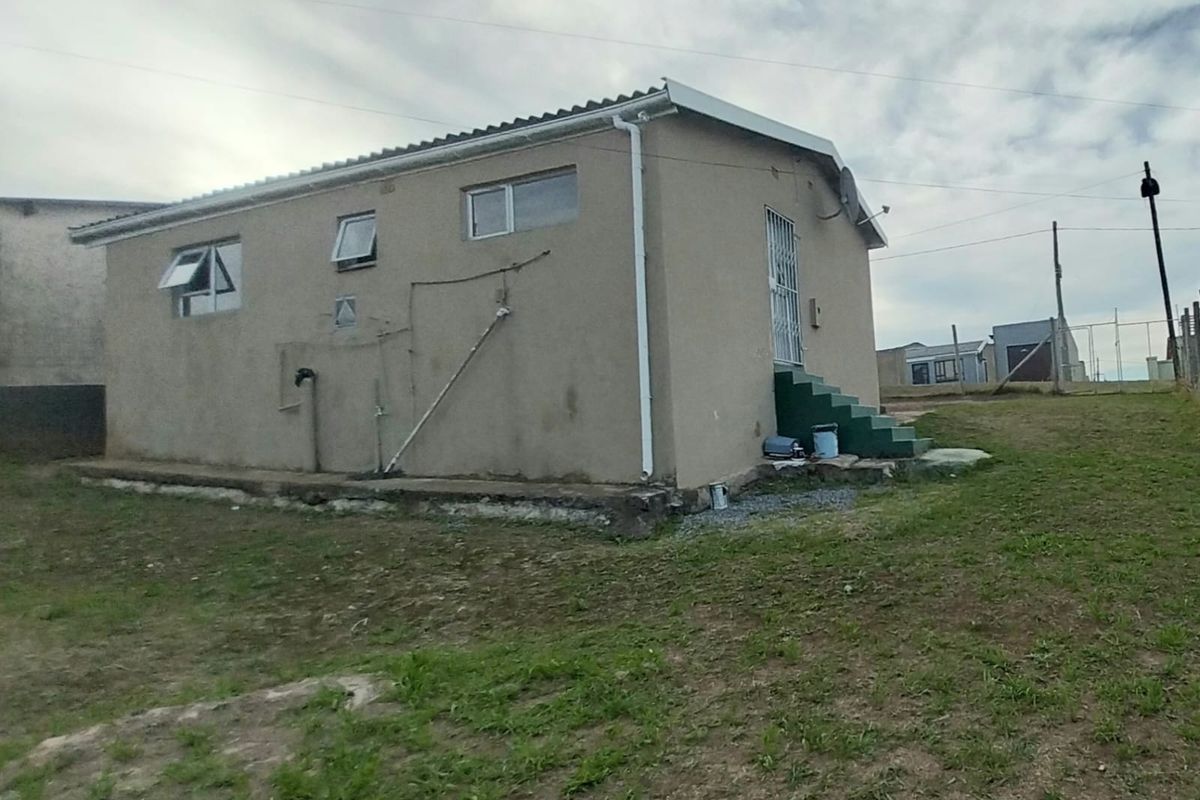 A side and rear view of a modest single-story house featuring plastered walls, a corrugated metal roof, and external plumbing fixtures.