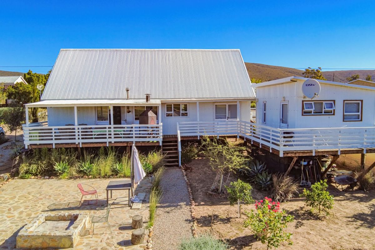 Rear view of a cottage-style house featuring a raised timber deck, corrugated metal roof, and a stone-paved patio area with a built-in fire pit.