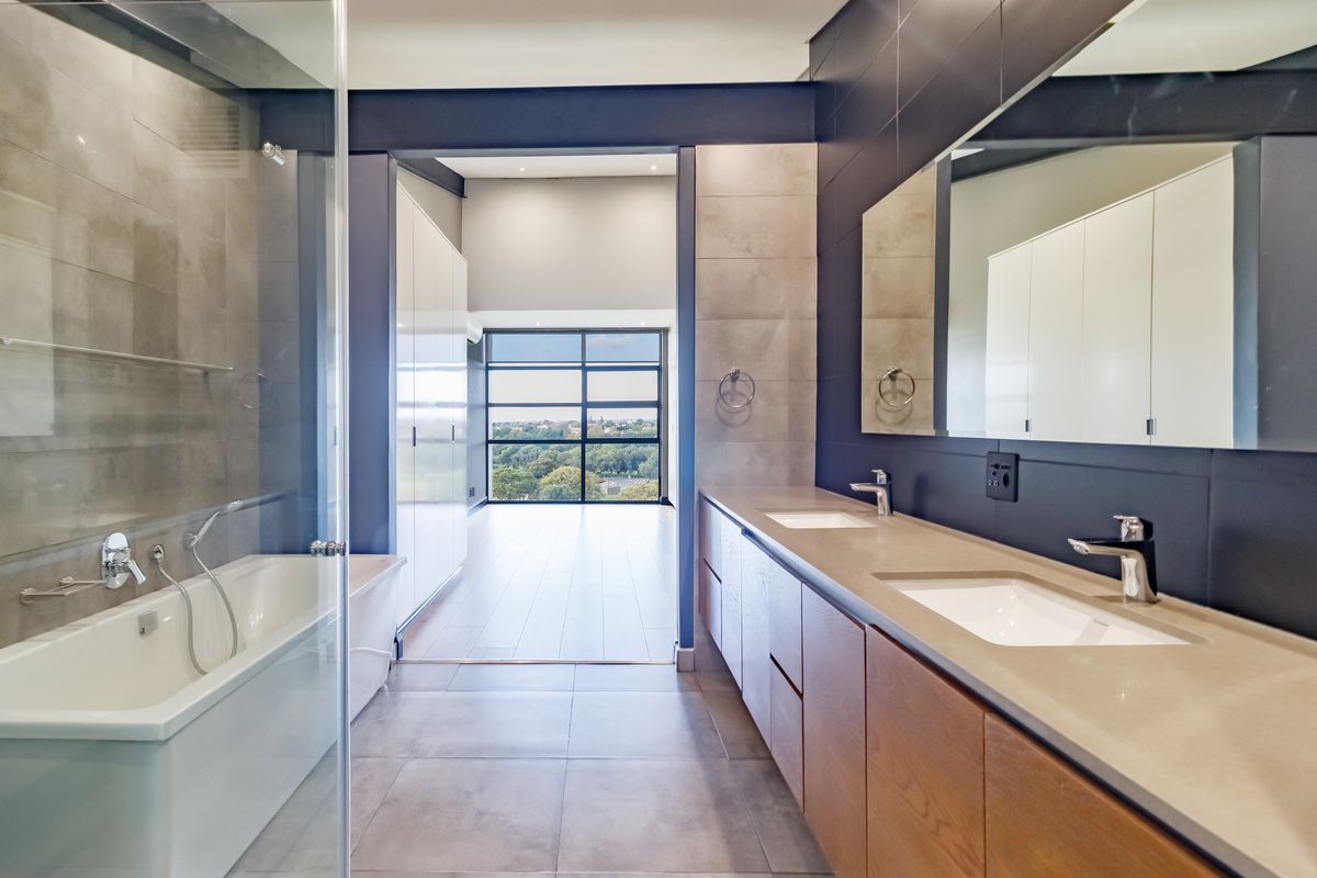 Modern bathroom featuring a double vanity with stone countertop, glass-enclosed bathtub, and large mirrors, leading into an open-plan living area with expansive views.