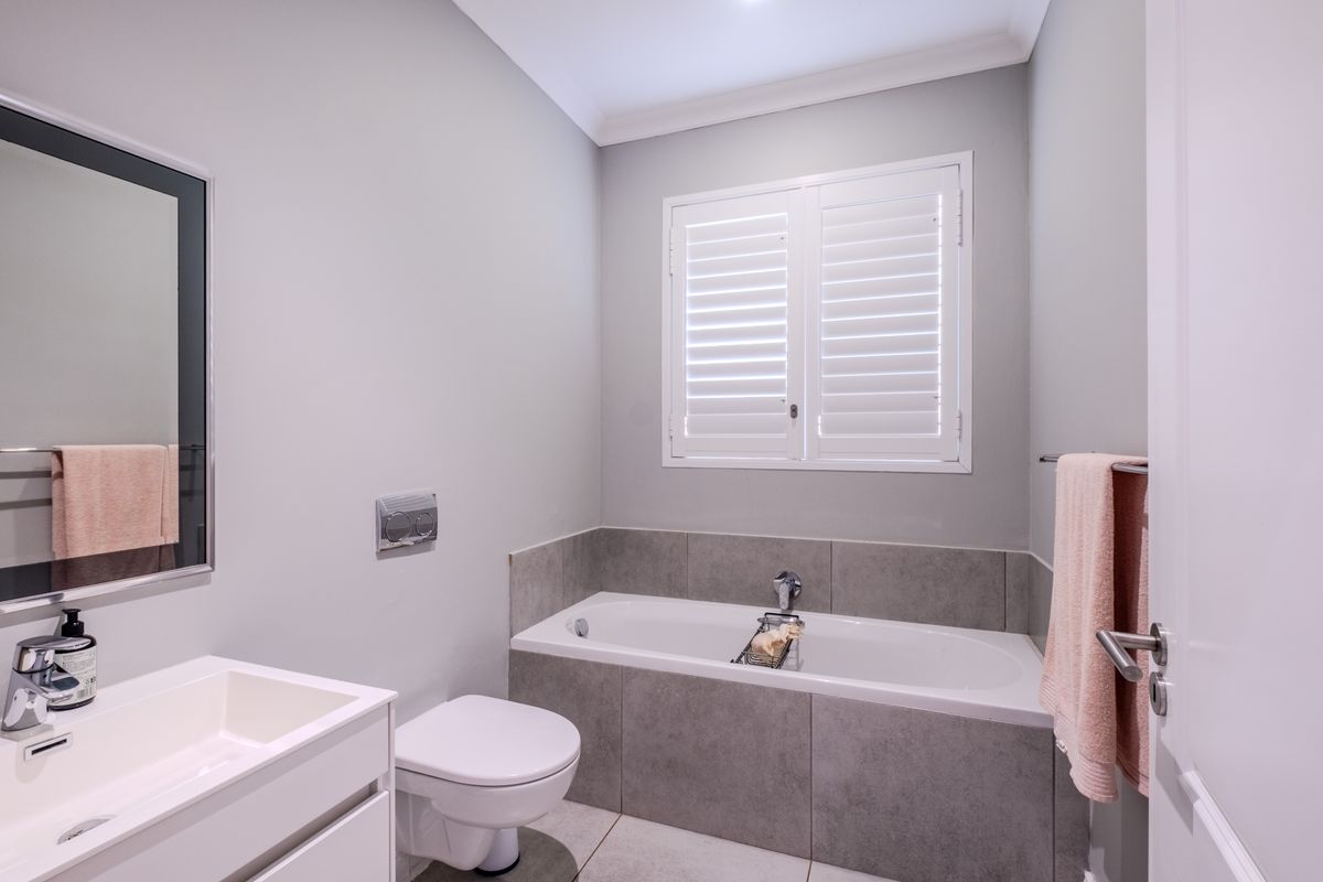 Modern bathroom featuring a built-in bathtub, wall-mounted toilet, sleek vanity, and plantation shutters, finished with contemporary grey tiling.