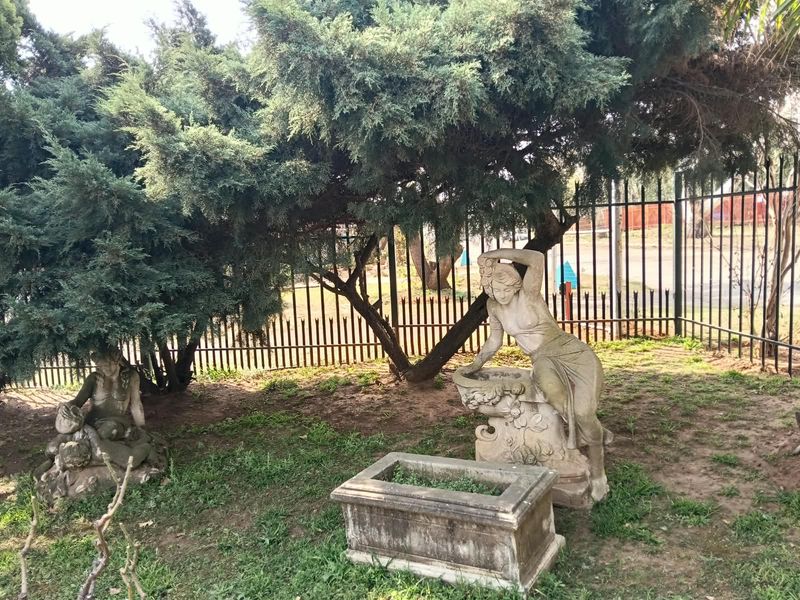 A landscaped garden area featuring mature trees, decorative stone statues, a stone planter, and secure metal perimeter fencing.