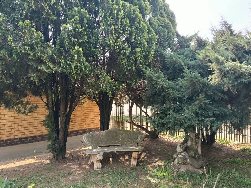 A landscaped garden area featuring mature trees, a brick boundary wall, and metal perimeter fencing.