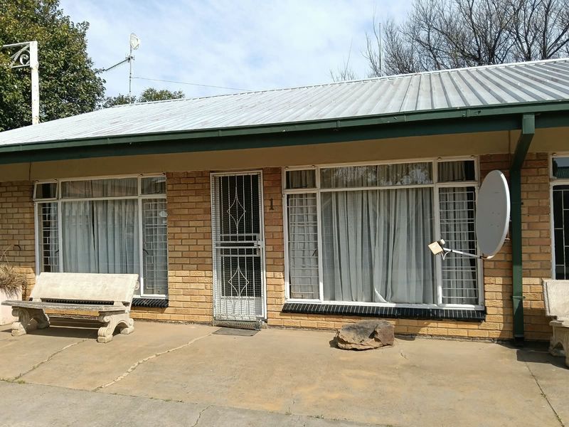 Front elevation of a brick apartment unit featuring a corrugated metal roof, security gates, and a paved patio area.