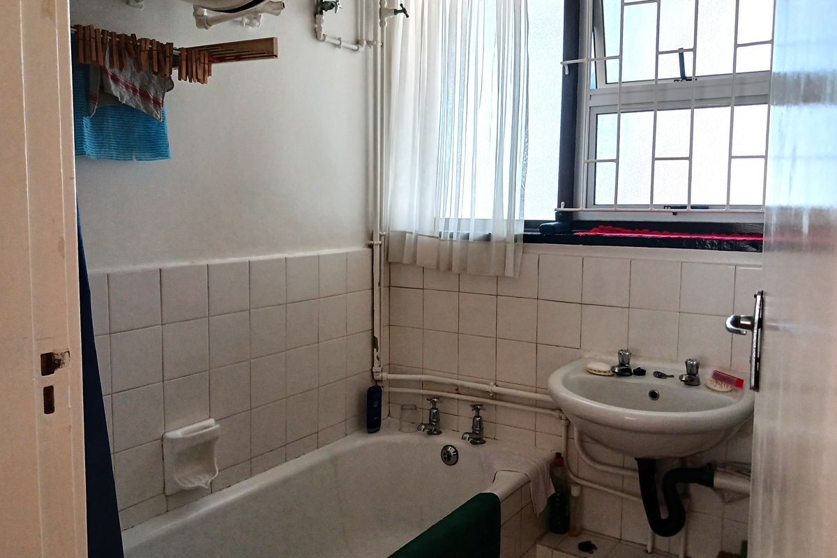 A functional bathroom featuring a standard bathtub, pedestal sink, and exposed plumbing, with tiled walls and a window with security bars.