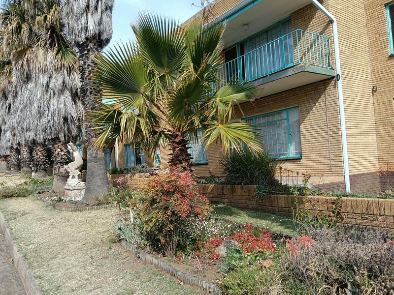 Exterior view of a brick apartment building featuring a balcony with metal railings and a landscaped front garden area.