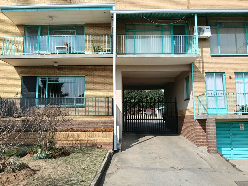 Front elevation of a brick apartment building featuring metal balcony railings, security gate access, and a concrete driveway.