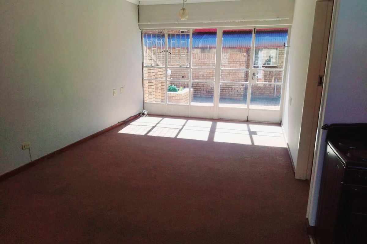 An empty, sunlit living room featuring large glass sliding doors with security bars and carpeted flooring, offering a blank canvas for renovation.