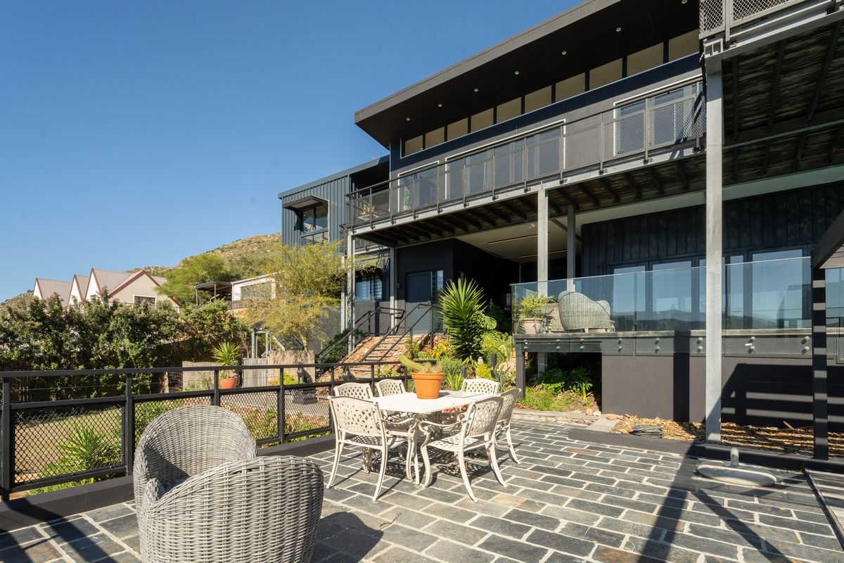 A modern multi-level rear exterior featuring a stone-paved patio, glass balustrades, and a contemporary dark-toned architectural facade with elevated decking.