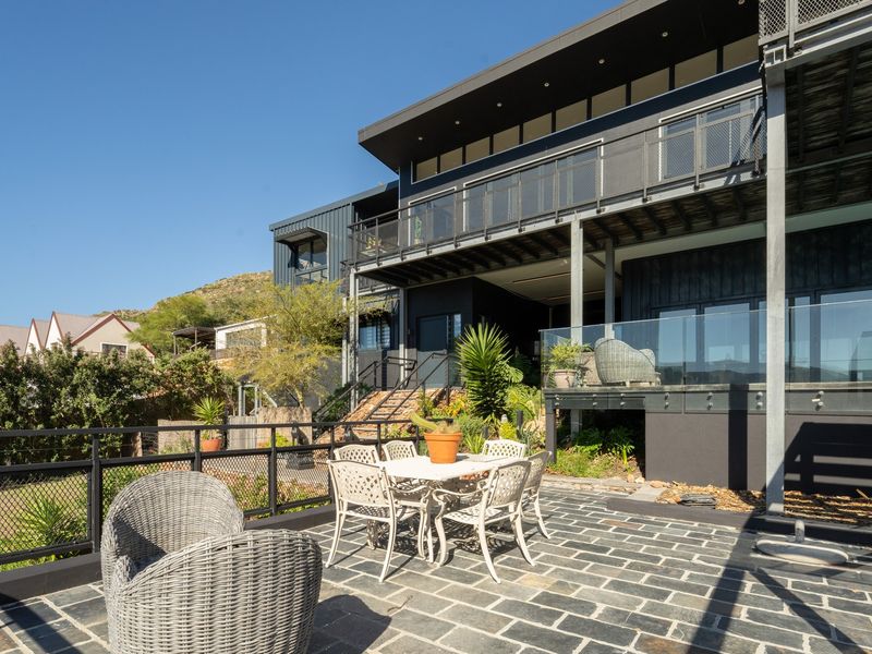 A modern multi-level rear exterior featuring a stone-paved patio, glass balustrades, and a contemporary dark-toned architectural facade with elevated decking.