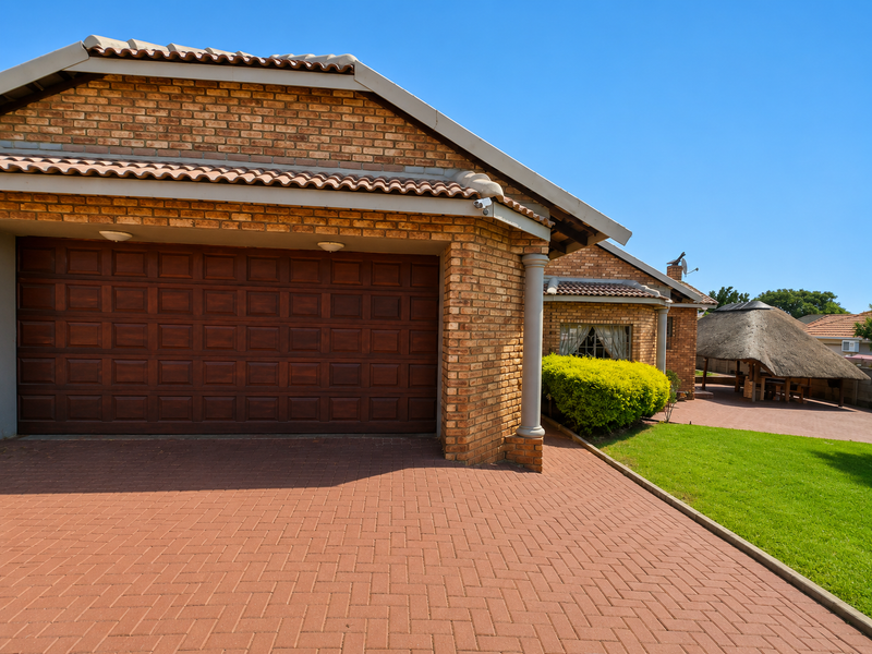 Front view of a brick house featuring a double garage, paved driveway, and a thatched lapa in the garden area.