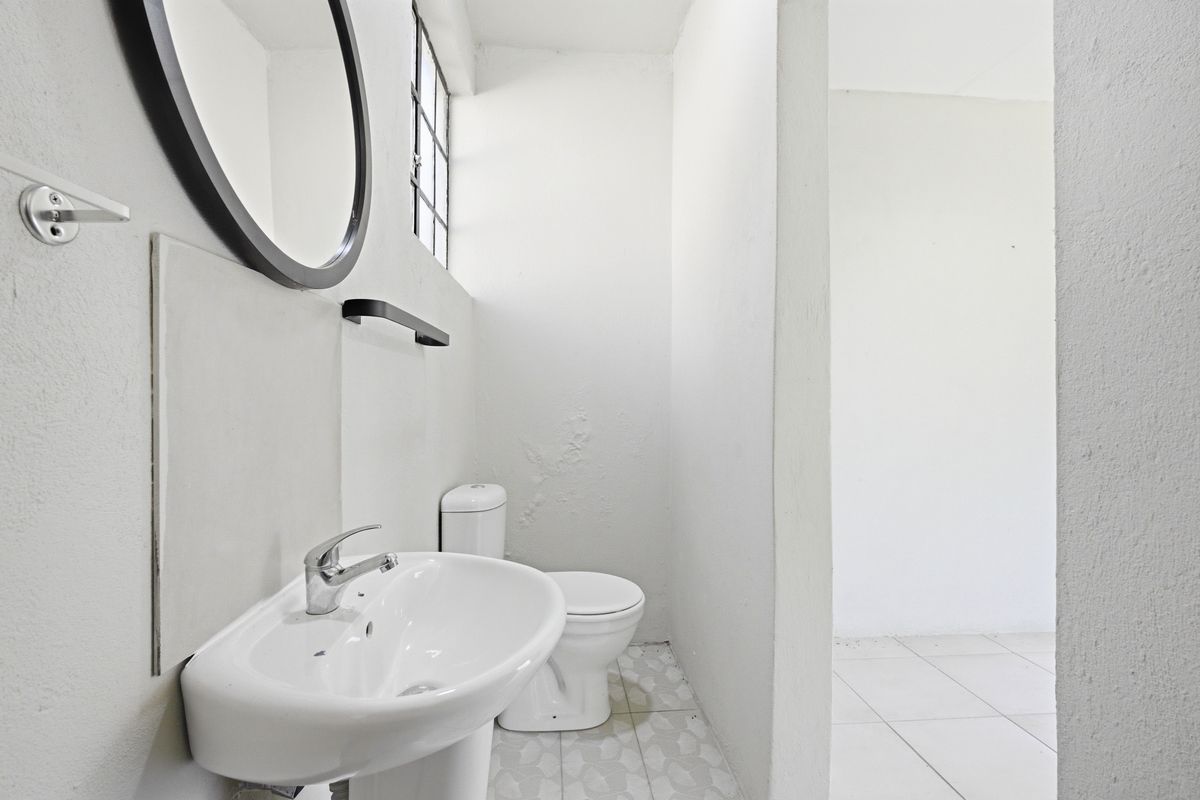 A compact bathroom featuring a pedestal sink, toilet, and small window, with tiled flooring and plain white walls.