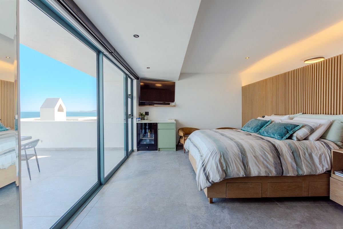 Modern bedroom featuring slatted timber wall paneling, recessed and cove lighting, tiled floors, and large sliding glass doors opening to a balcony with sea views.