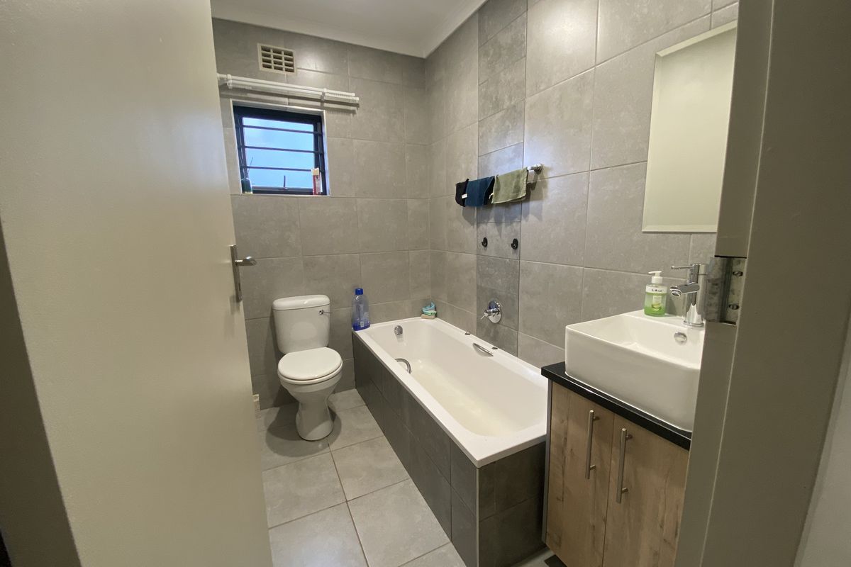 A modern bathroom featuring grey wall and floor tiles, a built-in bathtub, a vessel sink with a wooden vanity cabinet, and a toilet.