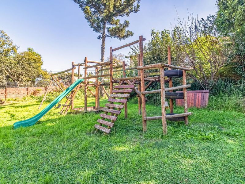 A spacious backyard garden featuring a large wooden jungle gym with a slide, set against a brick boundary wall and mature trees.