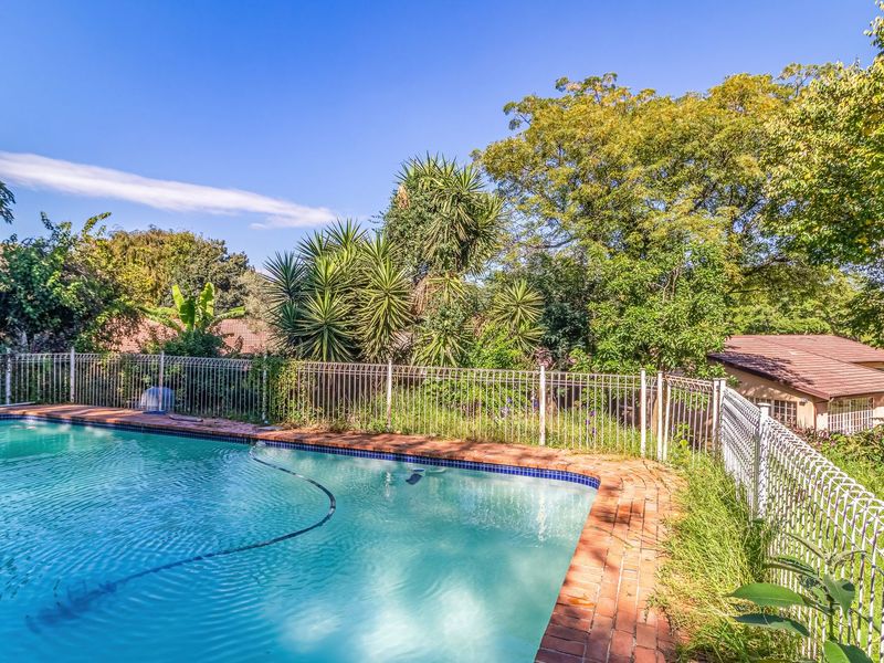 Outdoor swimming pool area featuring brick coping and metal security fencing, surrounded by established garden greenery.