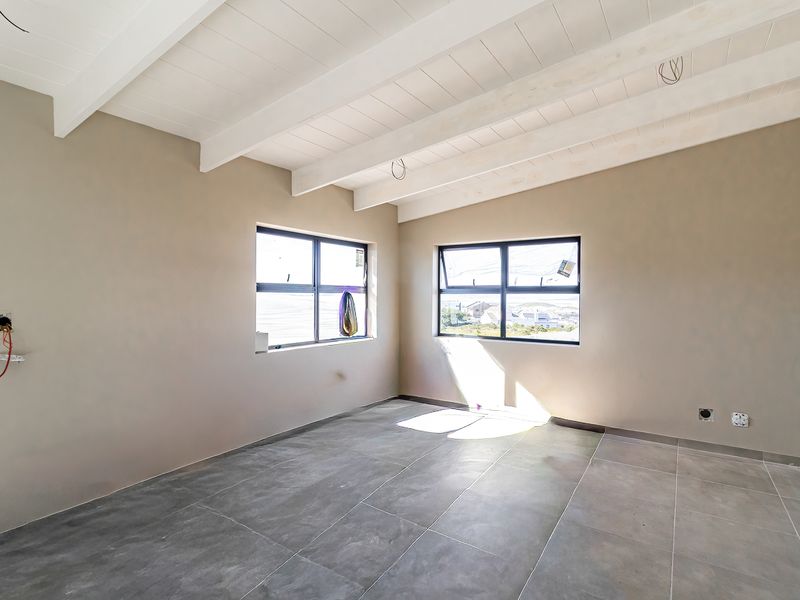 A bright, empty bedroom featuring exposed white timber ceiling beams, large aluminium windows, and modern grey floor tiles, currently awaiting final electrical finishing.