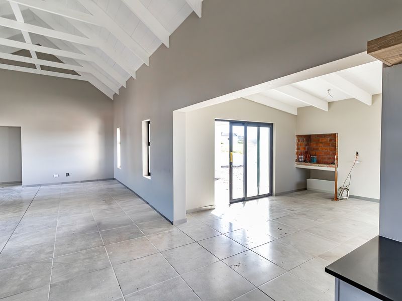 Spacious open-plan living area featuring vaulted ceilings with exposed rafters, large format floor tiles, and a built-in brick fireplace with direct outdoor access.
