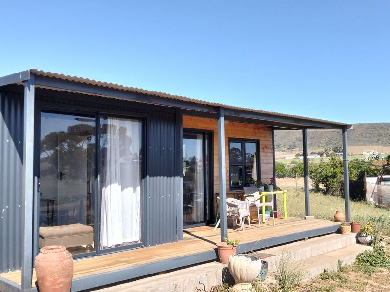 A modern modular-style home featuring a covered timber deck, corrugated metal exterior, and large sliding glass doors overlooking the surrounding landscape.