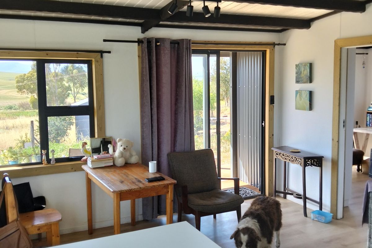 A bright living area featuring exposed timber ceiling beams, laminate flooring, and large windows with natural light and garden views.