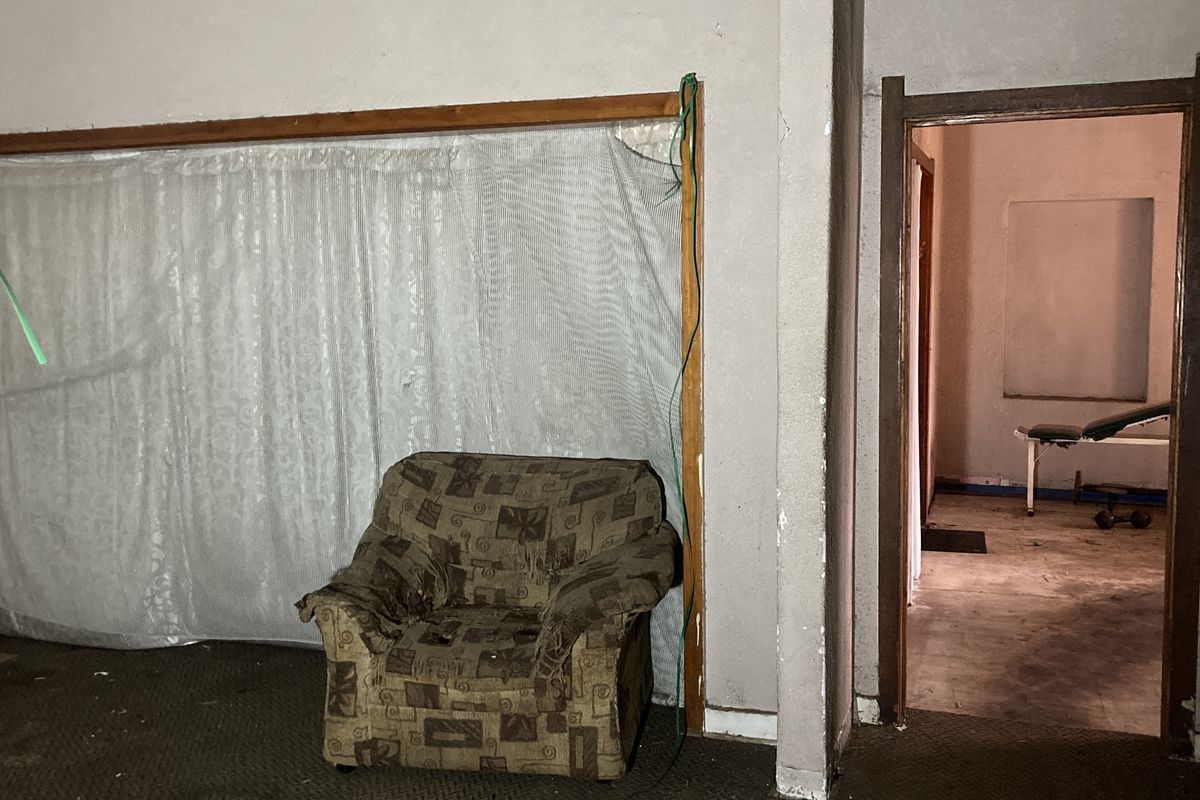 A living area in poor condition requiring significant renovation, featuring worn carpeting and damaged wall finishes, with an adjacent room visible through the doorway.