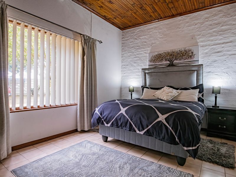 A well-maintained bedroom featuring a rustic exposed timber ceiling, textured painted brick wall, and neutral tiled flooring with natural light from large windows.