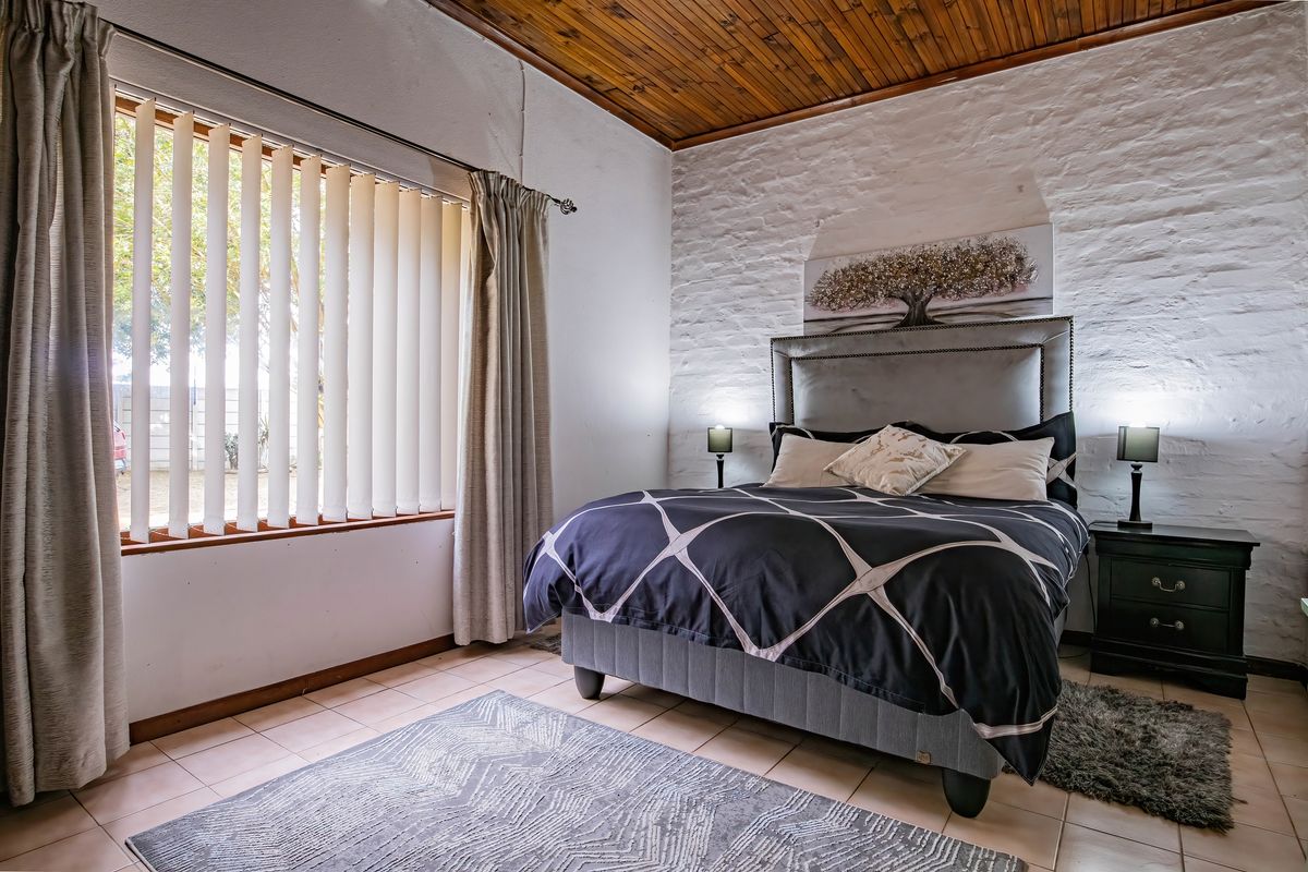 A well-maintained bedroom featuring a rustic exposed timber ceiling, textured painted brick wall, and neutral tiled flooring with natural light from large windows.