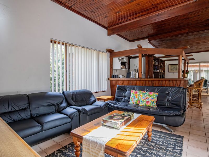 Open plan living area featuring a built-in timber bar, exposed wooden ceiling beams, and ceramic tiled flooring with natural light through vertical blinds.