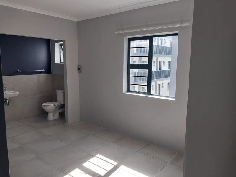 A bright, empty bedroom featuring neutral tiled flooring, a modern en-suite bathroom area with a wall-mounted basin, and large windows with security bars.