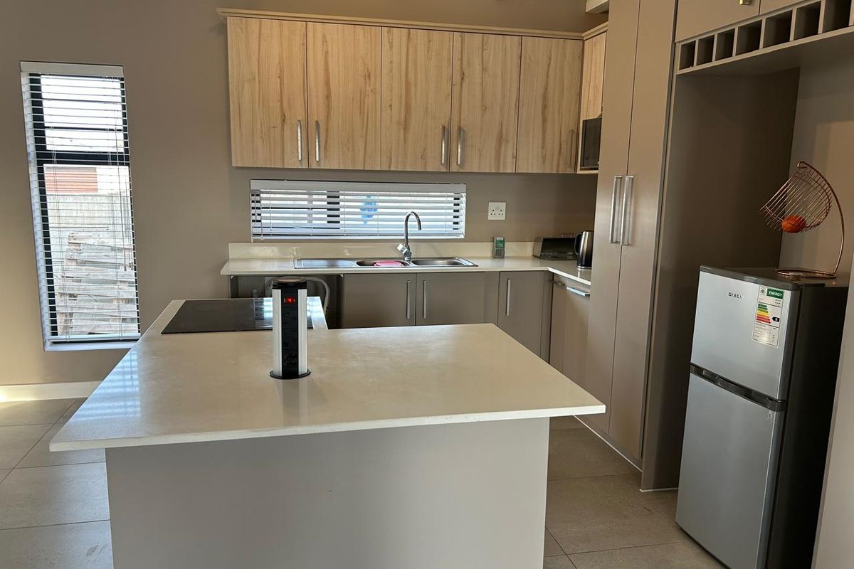 Modern kitchen featuring light wood-tone cabinetry, stone countertops, a central island with a built-in hob, and tiled flooring with exposed ceiling beams.