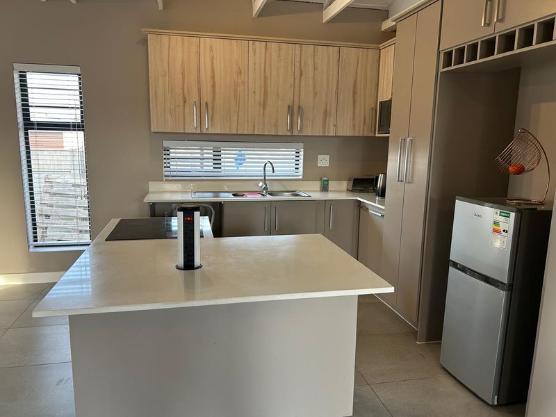 Modern kitchen featuring light wood-tone cabinetry, stone countertops, a central island with a built-in hob, and tiled flooring with exposed ceiling beams.