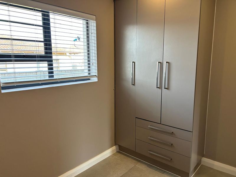 A clean, neutral bedroom featuring a modern built-in wardrobe with integrated drawers and a window with fitted venetian blinds.