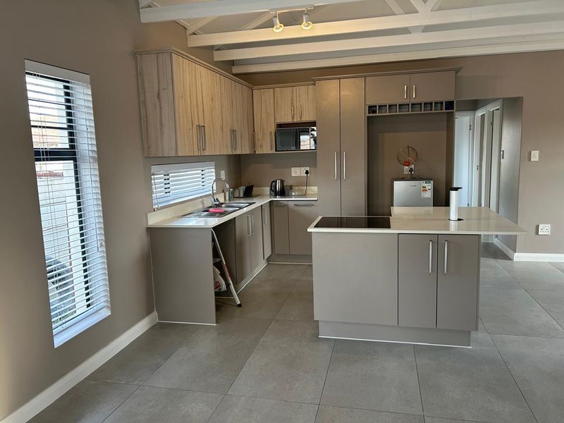 Modern kitchen featuring light wood and grey cabinetry, stone countertops, a central island with an electric hob, and exposed white ceiling beams.
