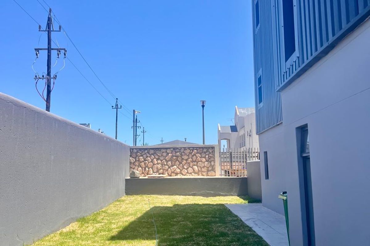 A private rear garden area featuring a manicured lawn, paved walkway, and modern metal-clad building exterior with high boundary walls.