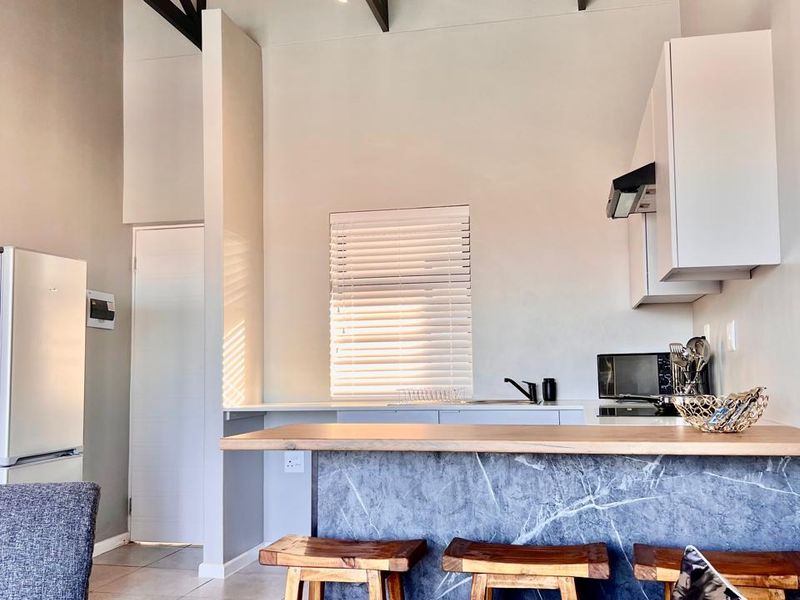 Modern open-plan kitchen and living area featuring high vaulted ceilings with exposed rafters, a stylish stone-look breakfast bar, and contemporary track lighting.