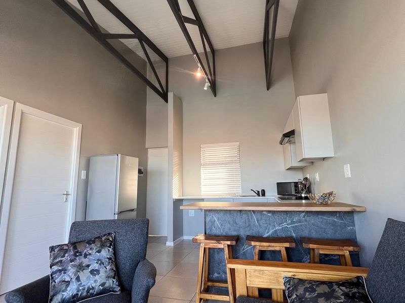 Open-plan living area featuring high ceilings with exposed steel trusses, tiled flooring, and a modern kitchen with a stone-look breakfast bar.
