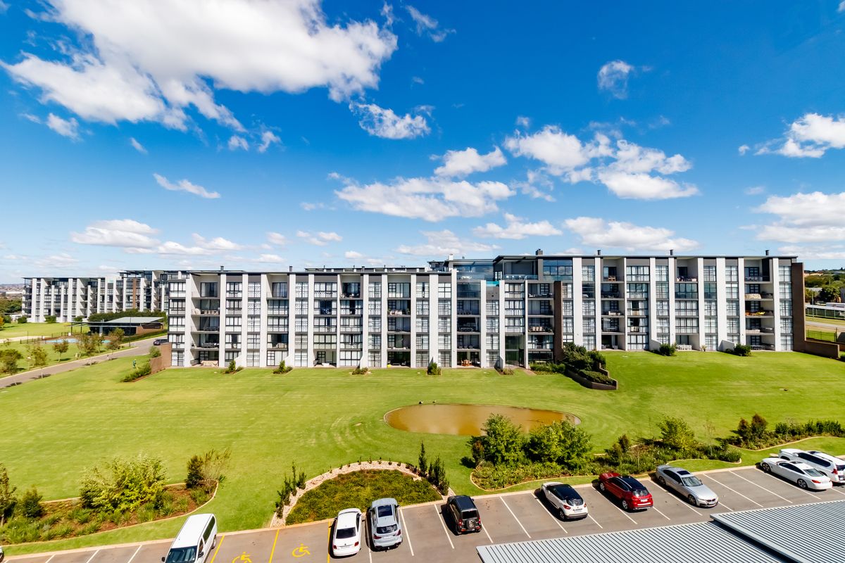 Aerial view of a modern multi-story apartment complex featuring extensive landscaped grounds, a central pond, and dedicated parking facilities.