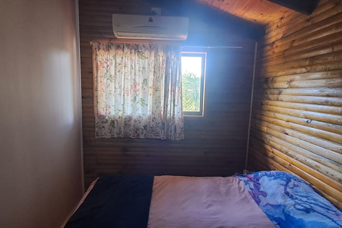 A cozy bedroom featuring rustic timber log walls, a wall-mounted split-system air conditioner, and natural light from a window.