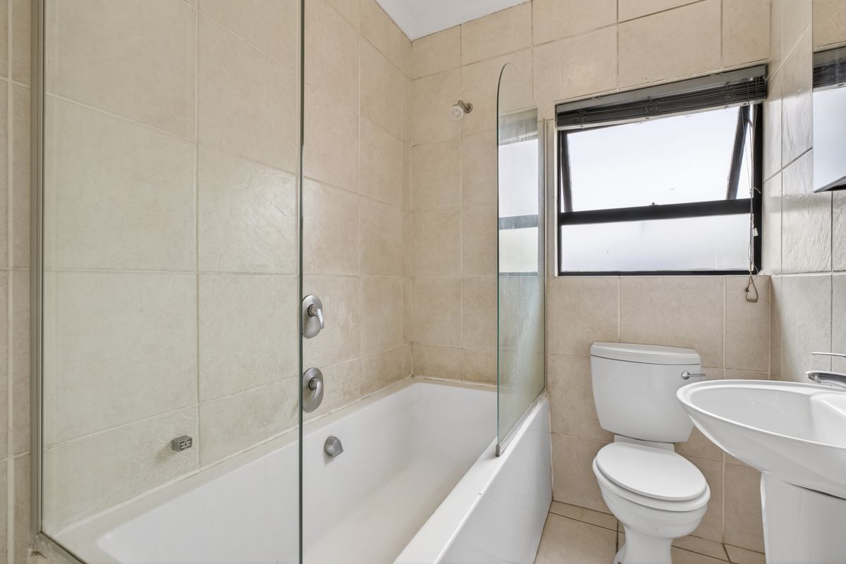 A functional bathroom featuring a bathtub with a glass shower screen, pedestal basin, and toilet, finished with neutral wall and floor tiling.
