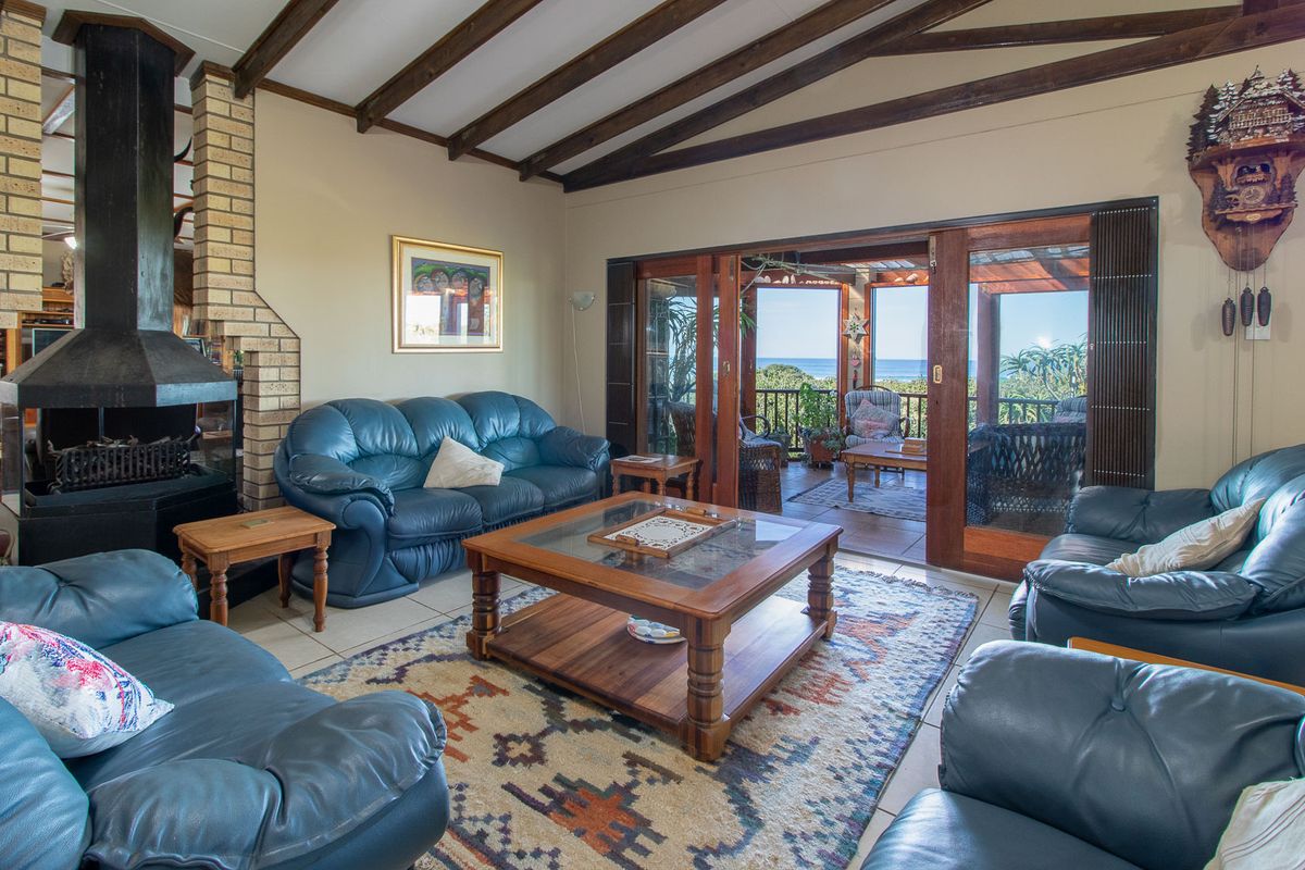 Spacious living room featuring exposed timber beams, a brick fireplace, and large sliding doors opening onto a patio with ocean views.