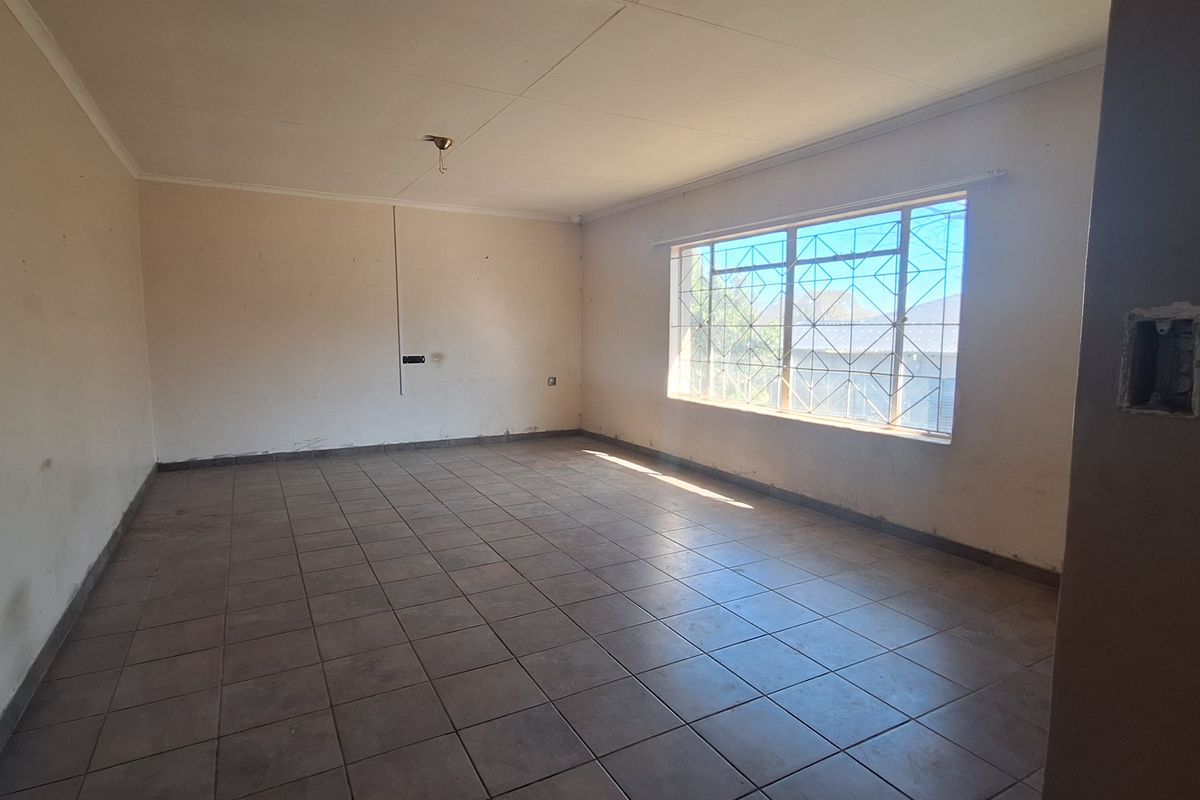 An empty, open-plan living area featuring tiled flooring and a large window with security bars, requiring cosmetic updates and minor electrical repairs.