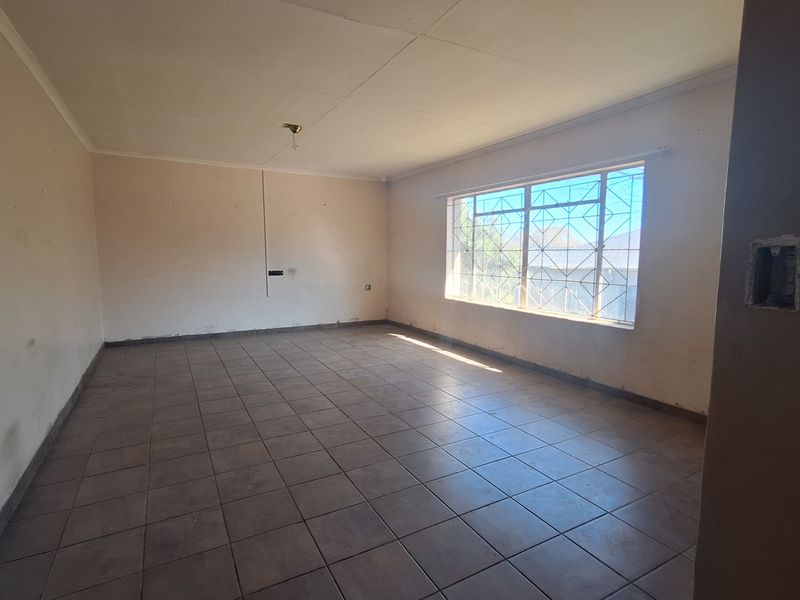 An empty, open-plan living area featuring tiled flooring and a large window with security bars, requiring cosmetic updates and minor electrical repairs.