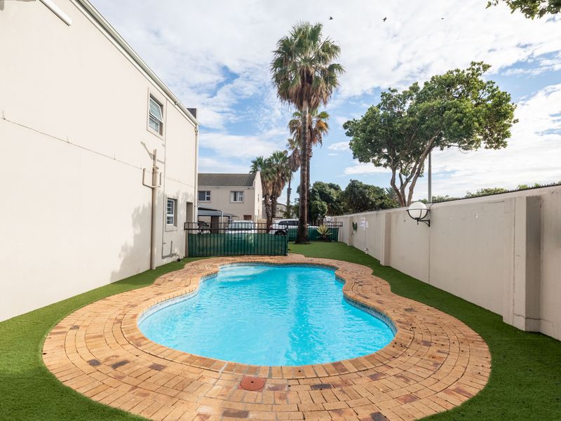 A private outdoor pool area featuring a kidney-shaped swimming pool with brick coping, surrounded by artificial turf and enclosed by high perimeter boundary walls.