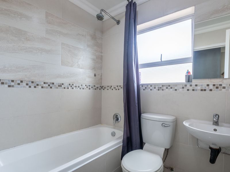 A functional bathroom featuring a bathtub with shower attachment, wall-mounted basin, and neutral tiled walls with a decorative mosaic border.