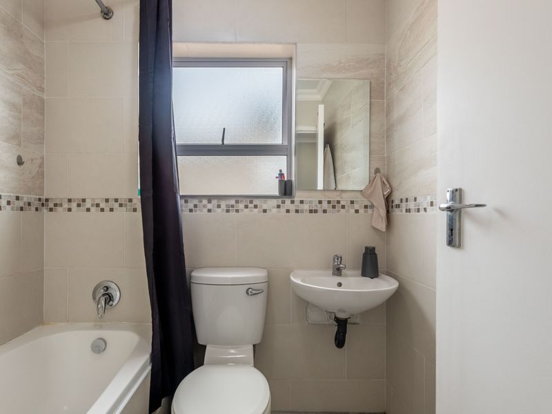 A compact, well-maintained bathroom featuring a bathtub with shower mixer, wall-mounted basin, and neutral tiled walls with a decorative mosaic border.