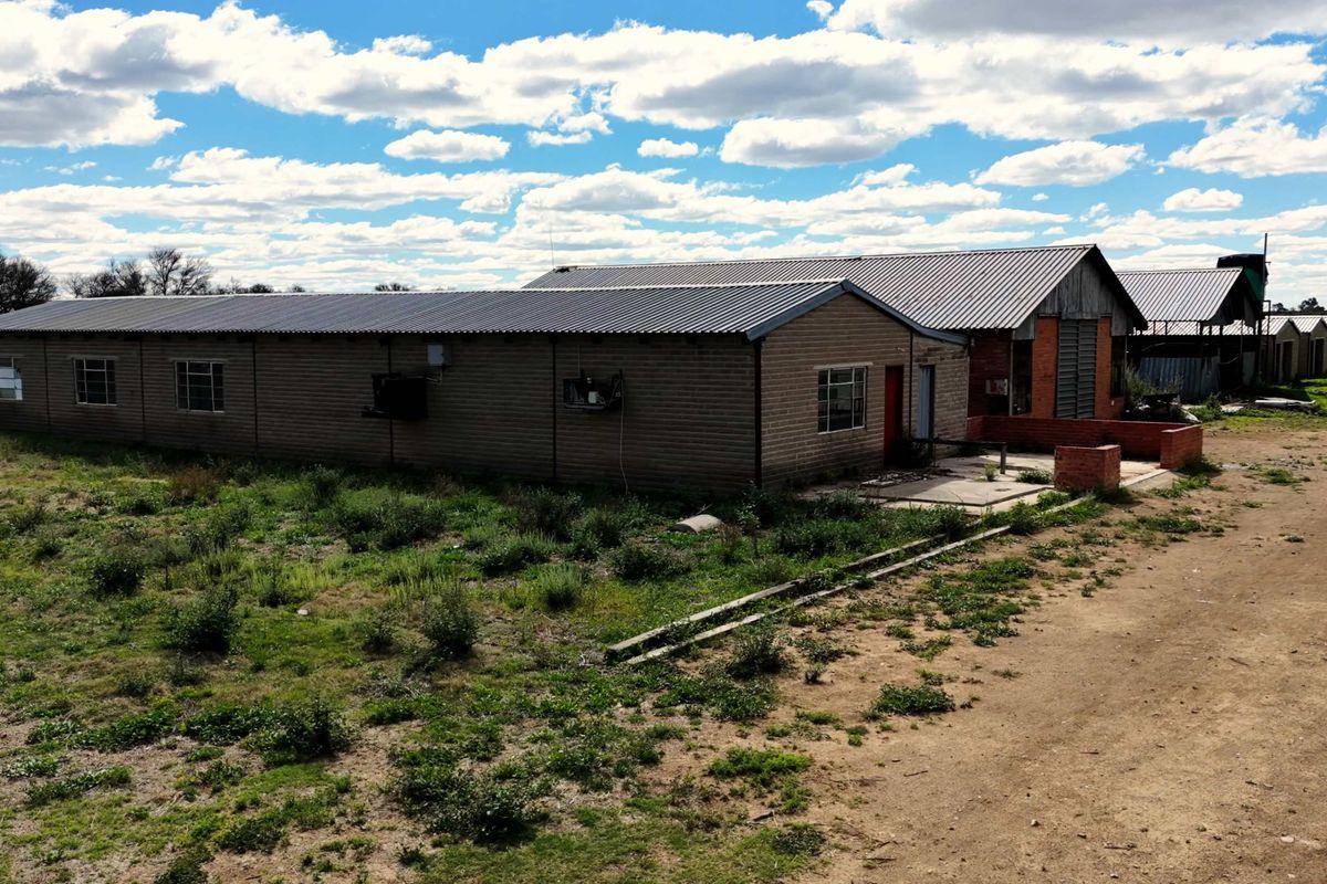 An aerial view of a farm building complex featuring long brick structures with corrugated metal roofing and surrounding unpaved land.