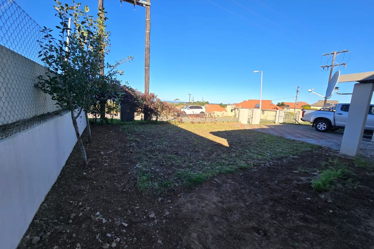 A rear yard area featuring a paved driveway, retaining wall, and chain-link fencing, with an open lawn space requiring landscaping attention.