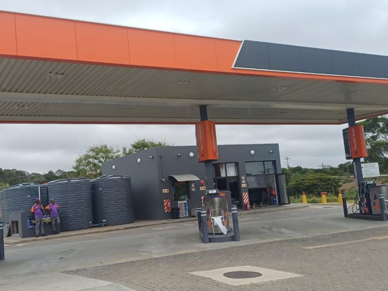 A commercial fuel station forecourt featuring a large canopy, multiple fuel pumps, and external water storage tanks on a paved site.