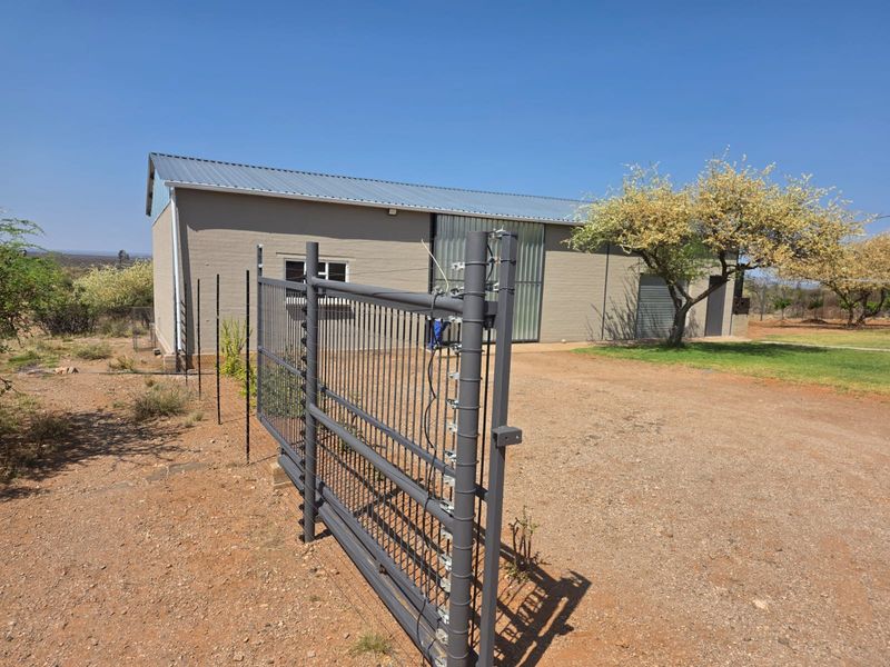 Exterior view of a farm building featuring a metal sliding gate, electric fencing, and a corrugated metal roof on a gravel driveway.